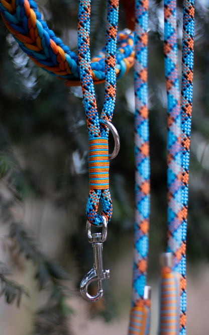 Na fotce vidíme krátké lanové vodítko - ručku neboli couračku, která se používá pokud chce člověk mít psa na krátko. Nechává se také na postroji pro rychlejší chycení psa na volno. Ručky jsou ručně pletené, mají zdobný oplet a mohou mít různé barvy, zde je také v pozadí lanové vodítko a paracordový obojek do barvy - modro - oranžová kombinace.